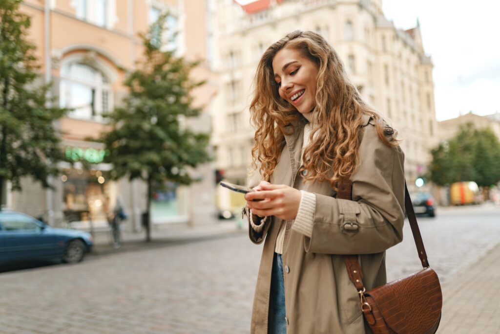 Smiling curly woman wearing warn coat walking down the street and using her phone